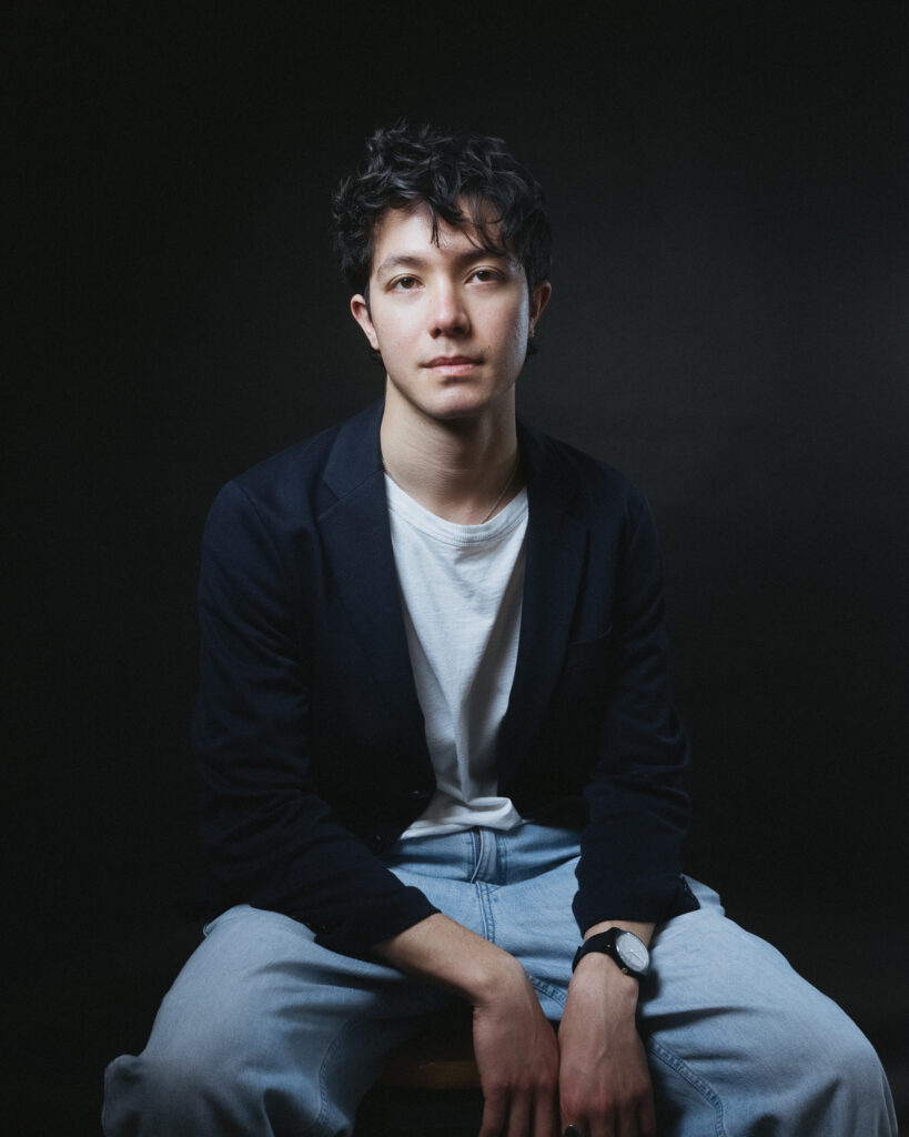 Photo of Hugh Rowlands (London-based) organist and pianist sitting against a black background. He wear blue jeans, a navy blazer, and a white t-shirt.
